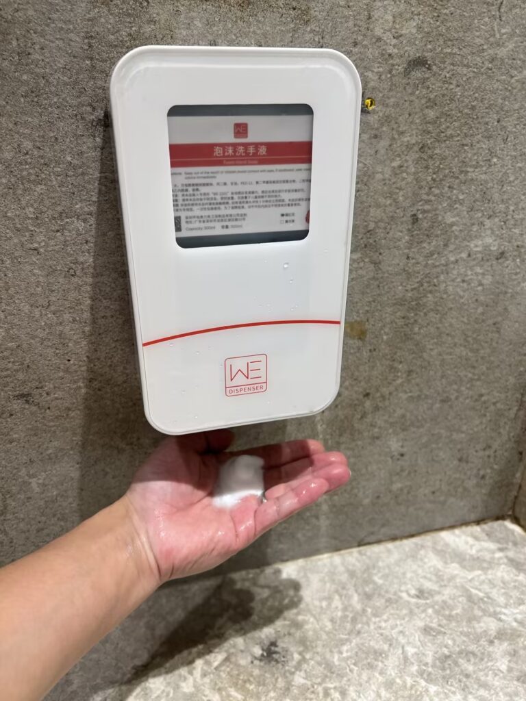 Hand receiving foam from a rectangular aolq we 2201 commercial wall mounted automatic soap dispenser with a viewing window and red markings installed on a concrete wall