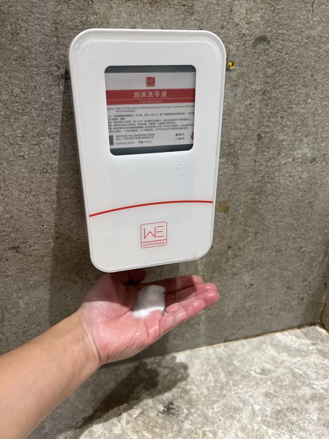 Hand receiving foam from a rectangular aolq we 2201 commercial wall mounted automatic soap dispenser with a viewing window and red markings installed on a concrete wall